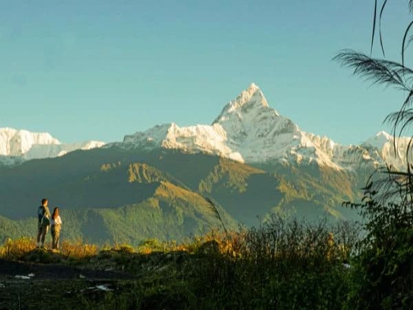 Two people standing on a hill with the snow-capped peak of Mount Machhapuchhre in the background at sunrise.
