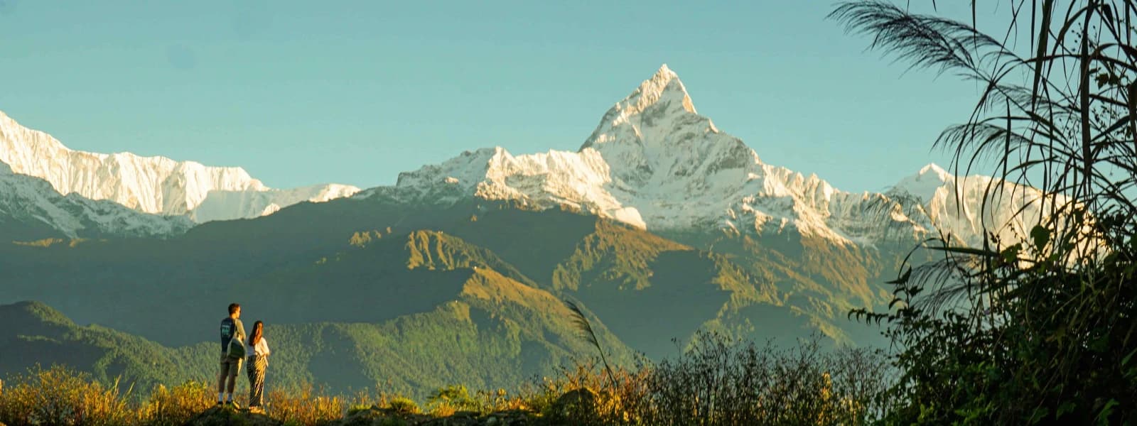 Two people standing on a hill with the snow-capped peak of Mount Machhapuchhre in the background at sunrise.